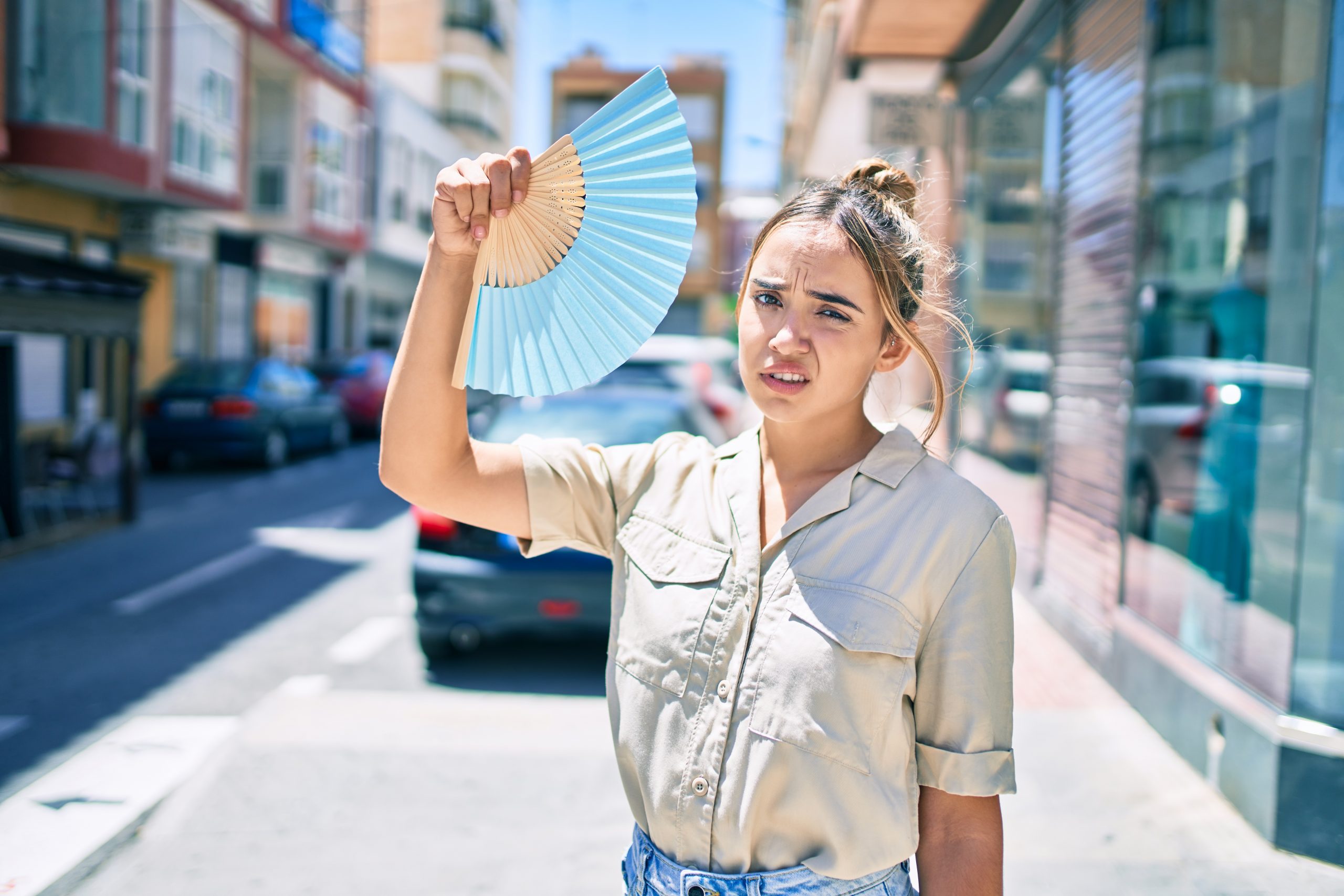 Young beautiful blonde caucasian woman smiling happy outdoors on a sunny day using handfan for hot weather